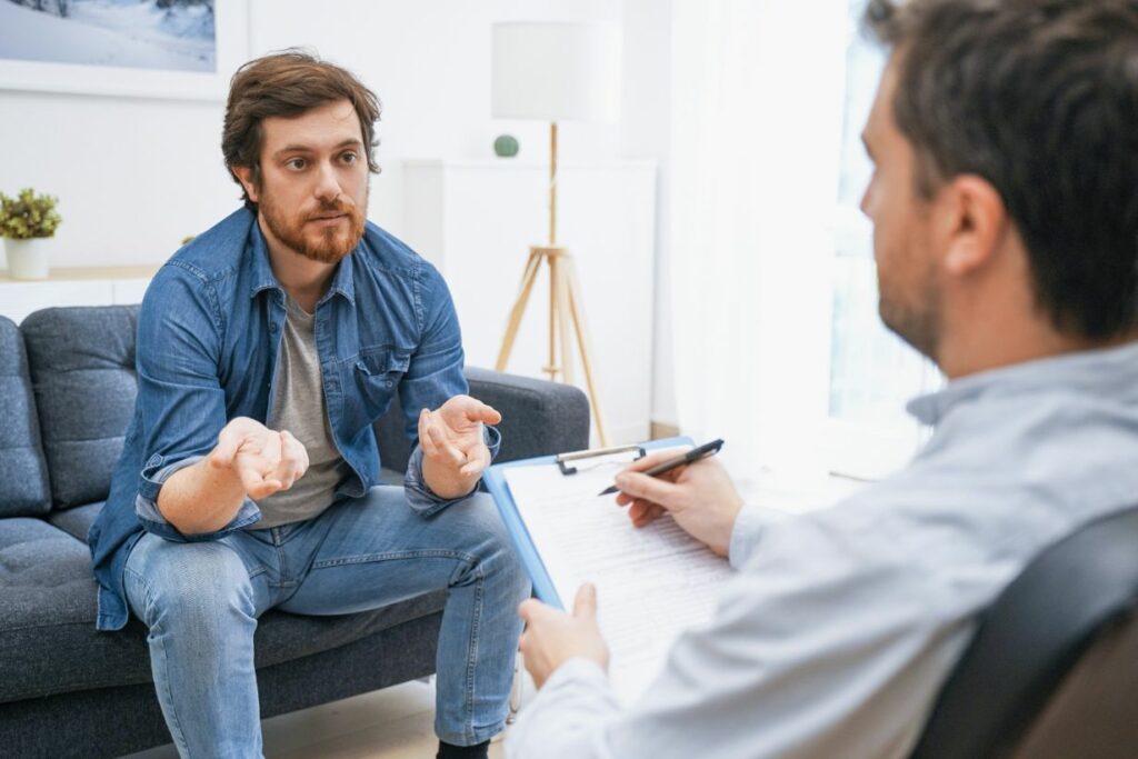 A man sits on a sofa, gesturing with his hands while speaking to another man holding a clipboard and pen during a conversation in a bright room.