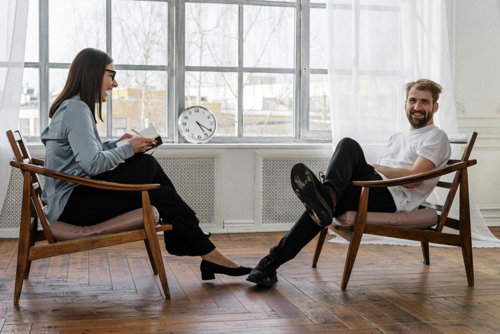 Two people sit facing each other in wooden chairs by a large window; one holds a notebook, the other is smiling with legs crossed. A clock is on the windowsill behind them.