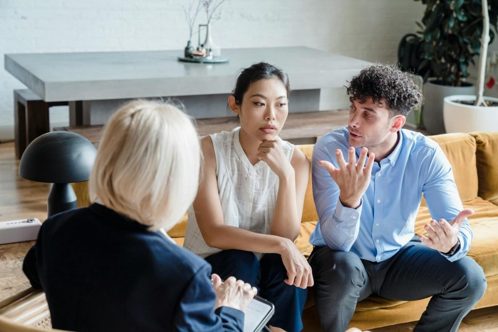 Three people sitting on a sofa in a living room. A woman on the left is writing on a clipboard while the two people on the right are engaged in conversation.