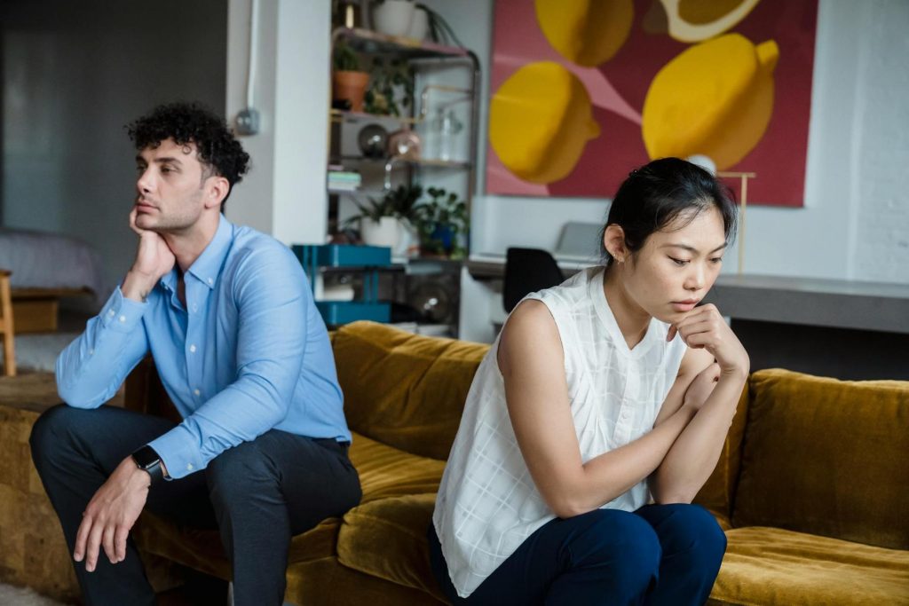 A man and a woman sit apart on a yellow couch, both looking thoughtful and distant, suggesting they might be experiencing tension or conflict.