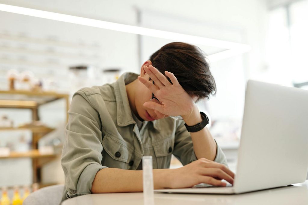 A person sitting at a table with a laptop, covering their face with one hand, possibly showing frustration or fatigue.