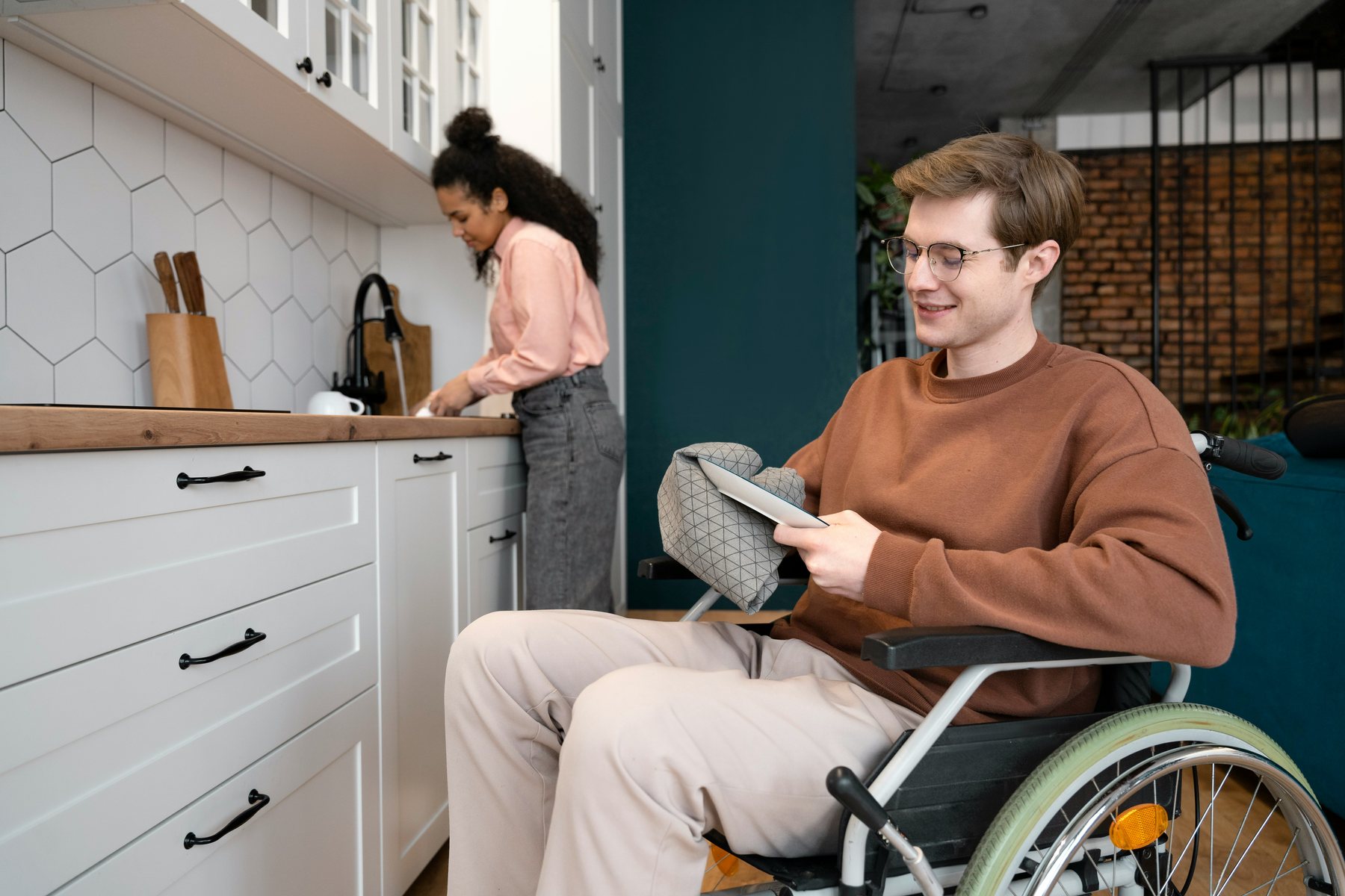 Young man in a wheelchair drying a plate in a bright kitchen while a woman washes up, practising daily living skills