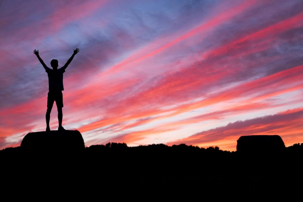 Silhouette of a person with raised arms on a hill against a colorful sunset sky with pink and orange clouds.