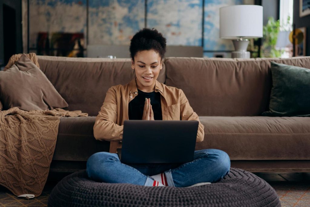 A person sits cross-legged on a cushion in front of a laptop, hands pressed together in a gesture of greeting or gratitude, with a sofa and home decor in the background.
