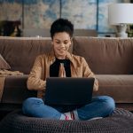 A person sits cross-legged on a cushion in front of a laptop, hands pressed together in a gesture of greeting or gratitude, with a sofa and home decor in the background.