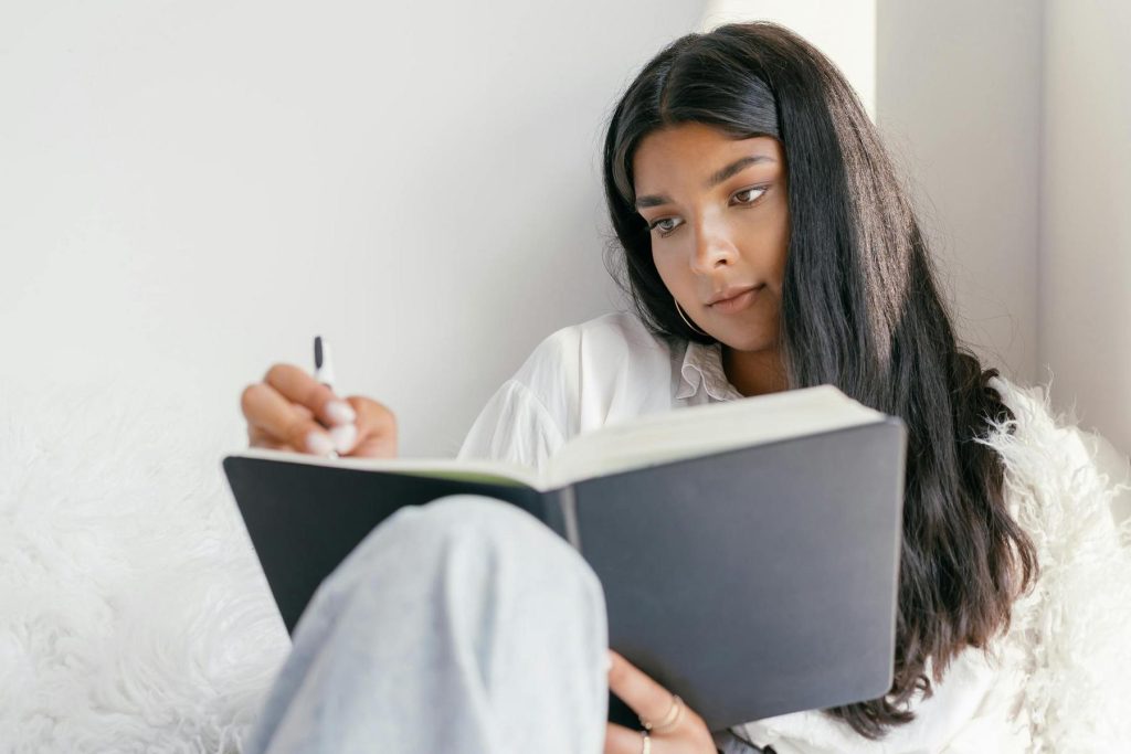 A person with long dark hair sits comfortably, writing in a large black notebook with a pen, looking focused and thoughtful.